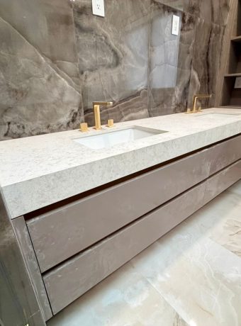 Modern bathroom vanity with custom white quartz countertop, integrated sinks, and full-height marble-look stone backsplash wall with brushed gold fixtures.
